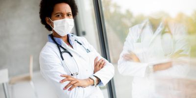 A Black female clinical investigator stands by a window with her arms crossed wearing a white lab coat and a stethoscope around her neck