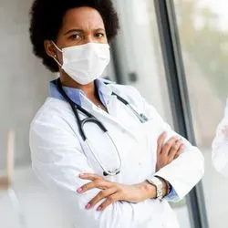 A Black female clinical investigator stands by a window with her arms crossed wearing a white lab coat and a stethoscope around her neck