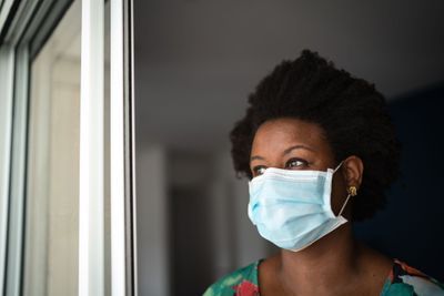 Photo of a black woman wearing a surgical mask looking out of a window