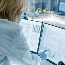 Image of a clinical lab professional wearing white lab coat and globes looking and pointing at quality control data on a computer monitor