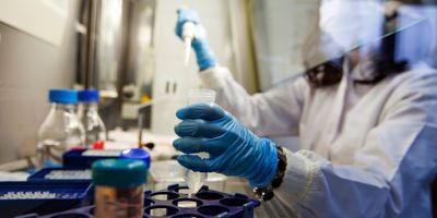 Photo of a clinical lab professional wearing PPE sitting at a biological safety cabinet pipetting COVID-19 dust samples