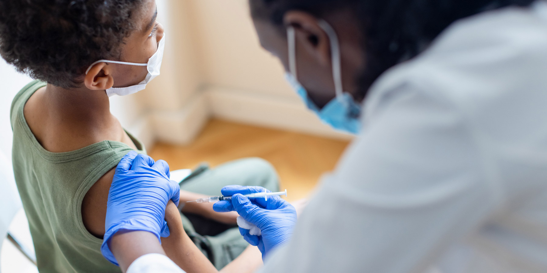 Photo of a Black clinician wearing PPE administering a vaccine to a Black five-year-old boy.