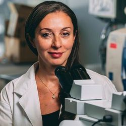 Photo of Dr. Gelareh Zadeh wearing a white lab coat sitting at a microscope at the Krembil Brain Institute at the University of Toronto.