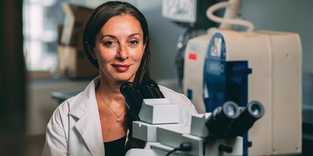 Photo of Dr. Gelareh Zadeh wearing a white lab coat sitting at a microscope at the Krembil Brain Institute at the University of Toronto.