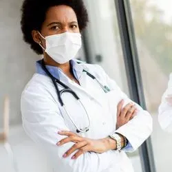 Photo of a Black woman clinical professional wearing a white lab coat and a surgical mask, standing with her arms crossed