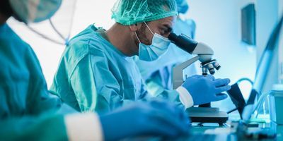 A laboratory professional wearing PPE looks at a slide on a microscope