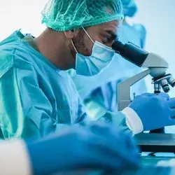 A laboratory professional wearing PPE looks at a slide on a microscope