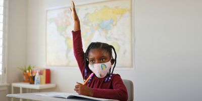 Child in school asking a question with hand up and wearing a mask