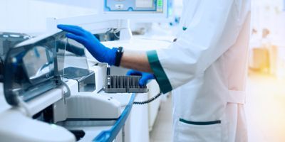 A laboratory worker wearing PPE handles automated hematology equipment
