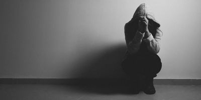 A person with a hood over their face and head in their hands crouching against a wall looking stressed