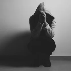 A person with a hood over their face and head in their hands crouching against a wall looking stressed
