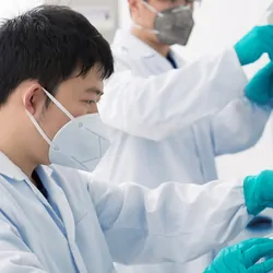 Medical laboratory workers in a lab wearing white lab coats, globes, and N95 masks handle SARS-CoV-2 samples
