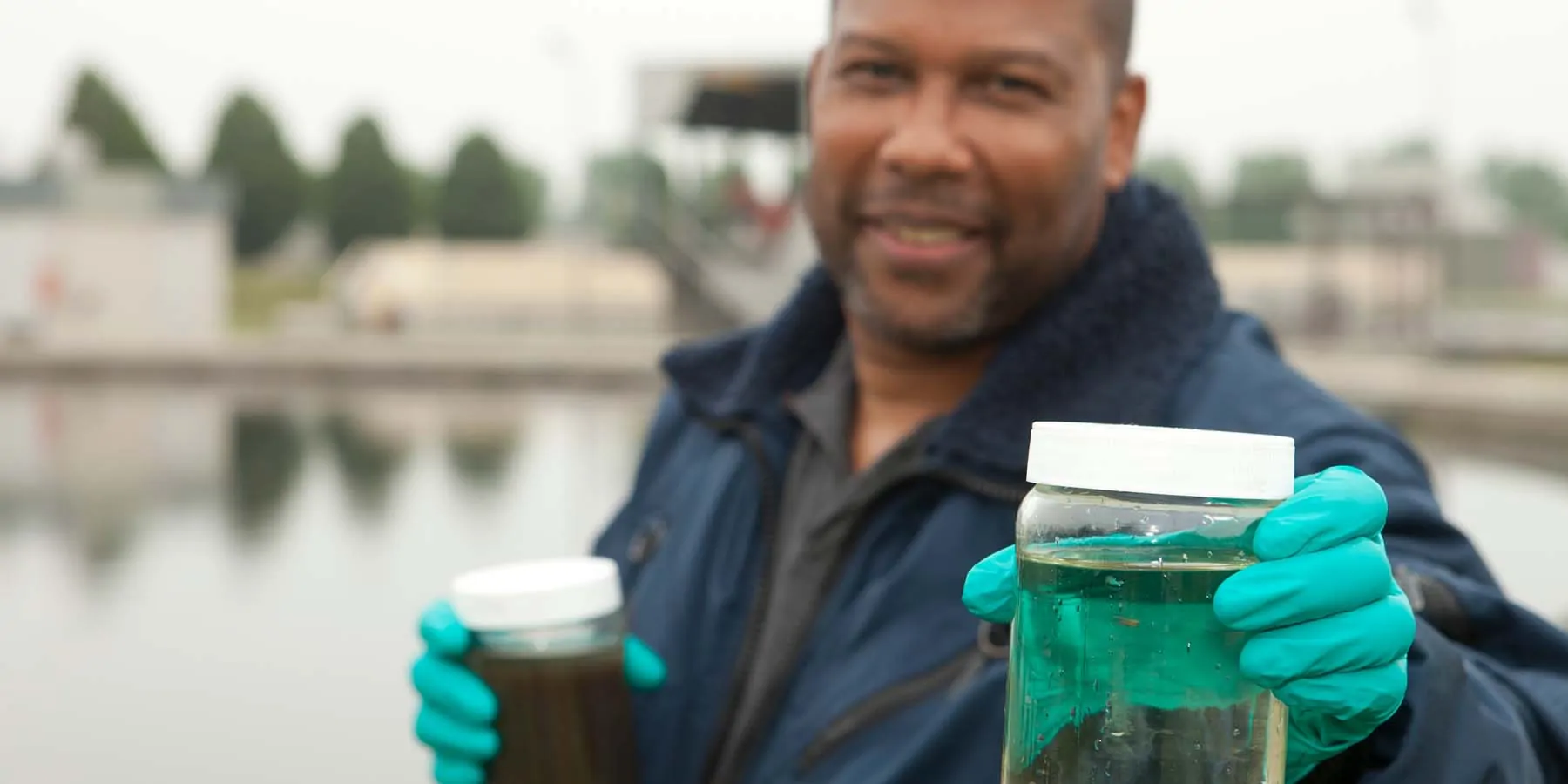 Photograph of a man working at a sewage treatment plant, holding COVID-19 water samples in each gloved hand. 