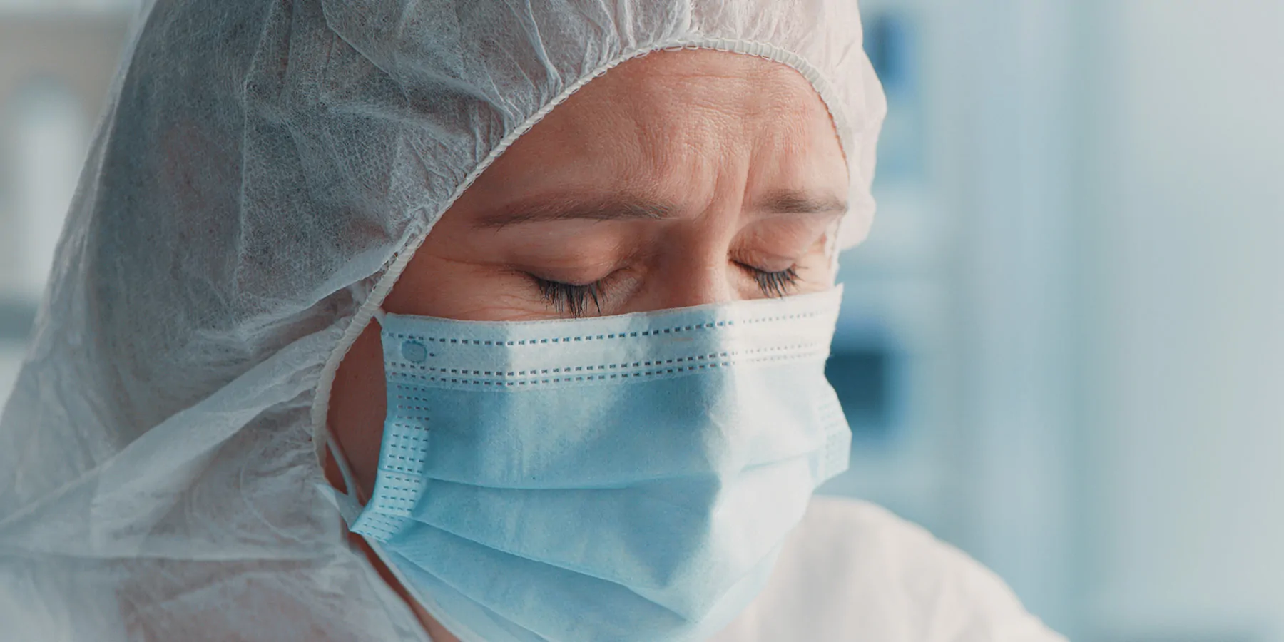 A clinical lab professional wearing a mask and PPE bonnet has their eyes closed, frowning.