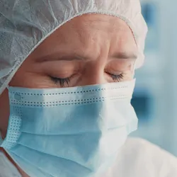 A clinical lab professional wearing a mask and PPE bonnet has their eyes closed, frowning.
