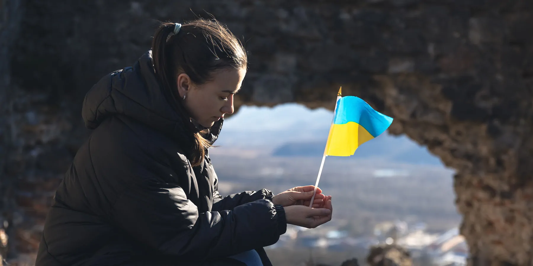 Photograph of a solemn young woman holding the flag of Ukraine as the country faces many barriers to health care. 