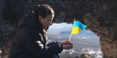 Photograph of a solemn young woman holding the flag of Ukraine as the country faces many barriers to health care. 