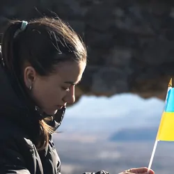 Photograph of a solemn young woman holding the flag of Ukraine as the country faces many barriers to health care.