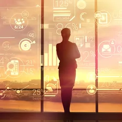 A clinical industry professional wearing a suit stands in front of a large window looking at a sunset.