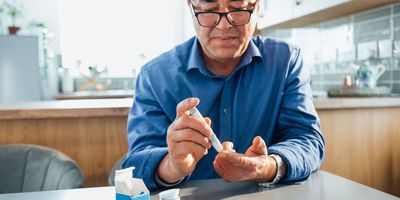 Asian man sitting at a dining table in his kitchen, he is pricking his finger using a glaucometer to test his blood sugar levels