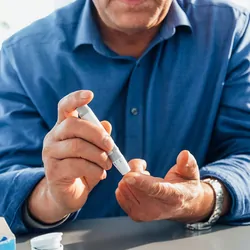 Asian man sitting at a dining table in his kitchen, he is pricking his finger using a glaucometer to test his blood sugar levels