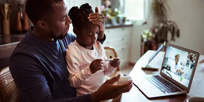 Photo of a Black man taking his daughter's temperature while on a telehealth call with a doctor.