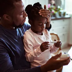Photo of a Black man taking his daughter's temperature while on a telehealth call with a doctor.