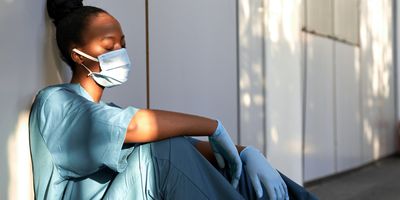 Tired exhausted scrub nurse wears face mask blue uniform gloves sits on hospital floor
