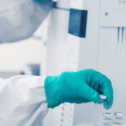 Photograph of clinical lab personnel picking up a vial of lyophilized reagent with their gloved hand, while another colleague performing a different experiment in the background.