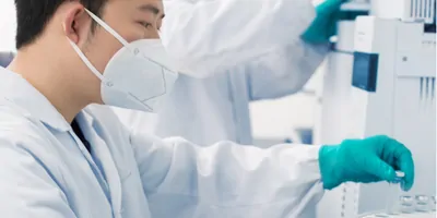 Photograph of clinical lab personnel picking up a vial of lyophilized reagents with his gloved hands, while another colleague works on another experiment in the background.
