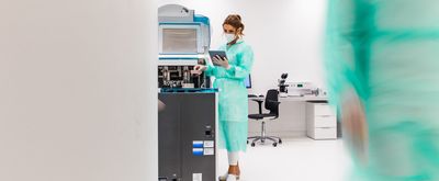 Woman in green gown and white mask with brown hair holds clipboard in a busy laboratory