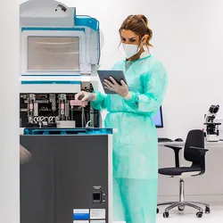 Woman in green gown and white mask with brown hair holds clipboard in a busy laboratory