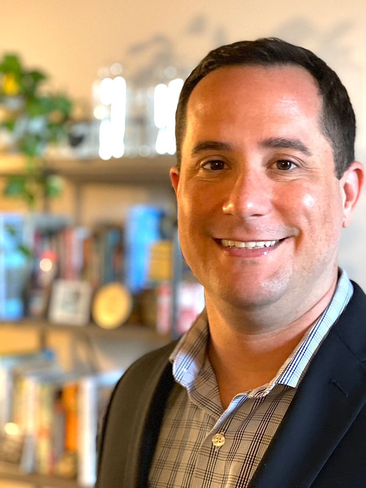 Man with black hair and brown eyes wearing black and white checked shirt and black blazer smiles at camera in professional headshot with bookshelves in background