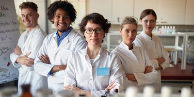 A group of medical laboratory students wearing white lab coats stand with their arms crossed in a clinical lab.