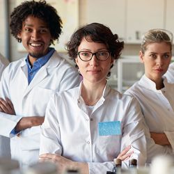 A group of medical laboratory students wearing white lab coats stand with their arms crossed in a clinical lab.
