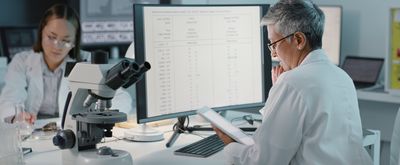 Women wearing white lab coats look at monitor of data with microscope sitting nearby.