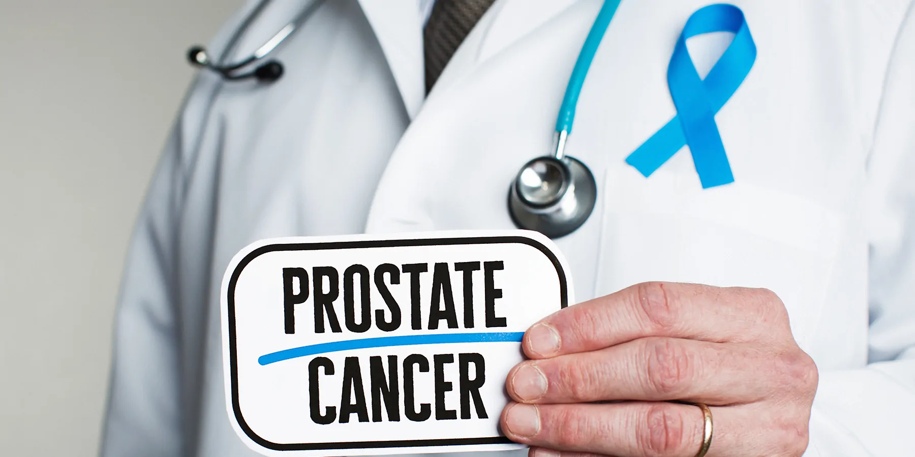 A person wearing a lab apron, blue ribbon, and stethoscope flashes a card with his left hand having the words prostate cancer.