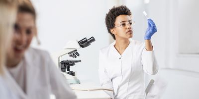 Lab personnel, seated near a microscope, examines immunohistochemistry slide with her gloved hand, prior to placing the cancer specimen under the microscope.