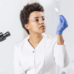 Lab personnel, seated near a microscope, examines immunohistochemistry slide with her gloved hand, prior to placing the cancer specimen under the microscope.
