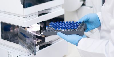 Close-up of a personnel's hands with gloves loading a tray of sample vials into a liquid chromatography machine.