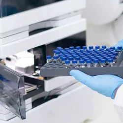 Close-up of a personnel's hands with gloves loading a tray of sample vials into a liquid chromatography machine.