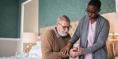A female personal caregiver helps a male senior patient stand up from the bed.
