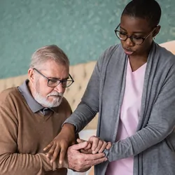 A female personal caregiver helps a male senior patient stand up from the bed.