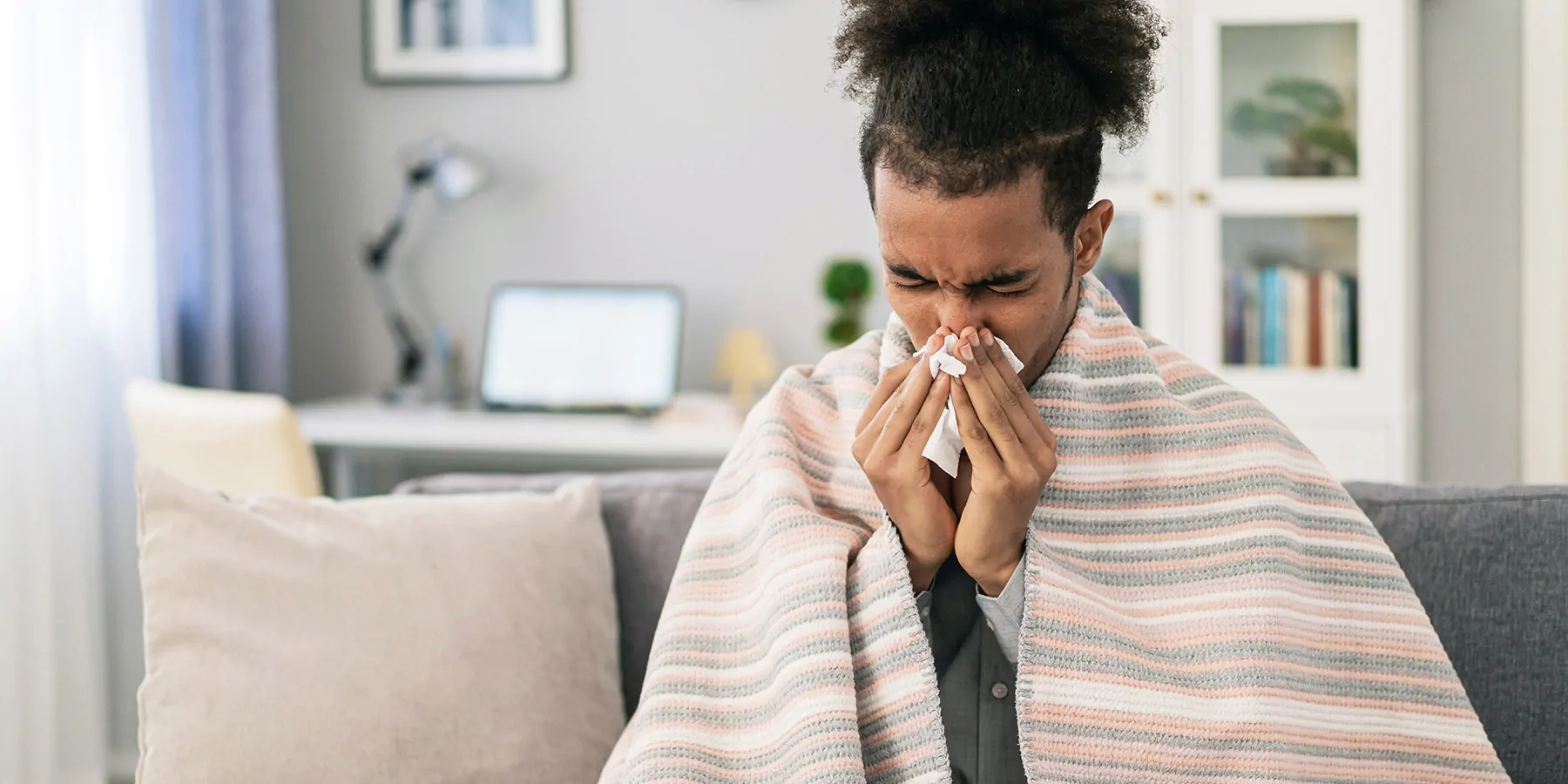 Sick young African-American man sitting on the sofa, covered with blanket at home and blowing his nose