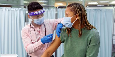 A young Black woman wearing a face mask sits in a medical clinic as a male doctor of Asian descent prepares to give her a COVID-19 vaccination injection in the upper arm.