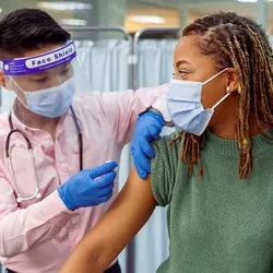 A young Black woman wearing a face mask sits in a medical clinic as a male doctor of Asian descent prepares to give her a COVID-19 vaccination injection in the upper arm.