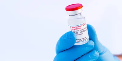 Close-up of a gloved hand holding a Moderna COVID-19 vaccine vial against a white background.