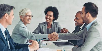 Businesswoman and businessman shaking hands across the table with more people in the background.