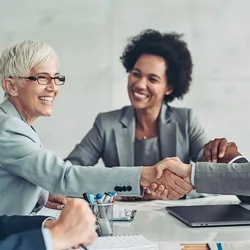 Businesswoman and businessman shaking hands across the table with more people in the background.