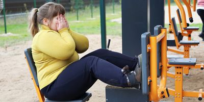 An overweight girl covers her face with hands cupped as she sits on workout equipment in an open area.